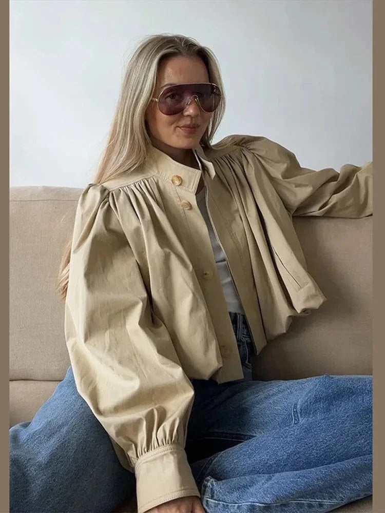 Woman wearing a beige jacket and sunglasses sitting on a couch.