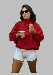 Woman in red jacket and white shorts holding a coffee cup and cookie in front of a wooden door.