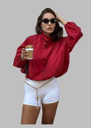 Woman in red jacket and white shorts holding a coffee cup against a beige wall.