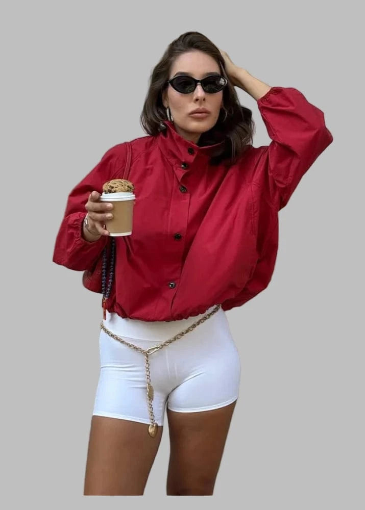 Woman in red jacket and white shorts holding a coffee cup against a beige wall.