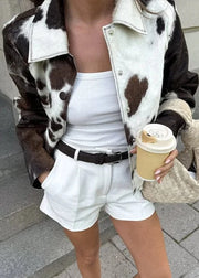 Person wearing a cow print jacket, white top, and shorts, holding a coffee cup and bag.