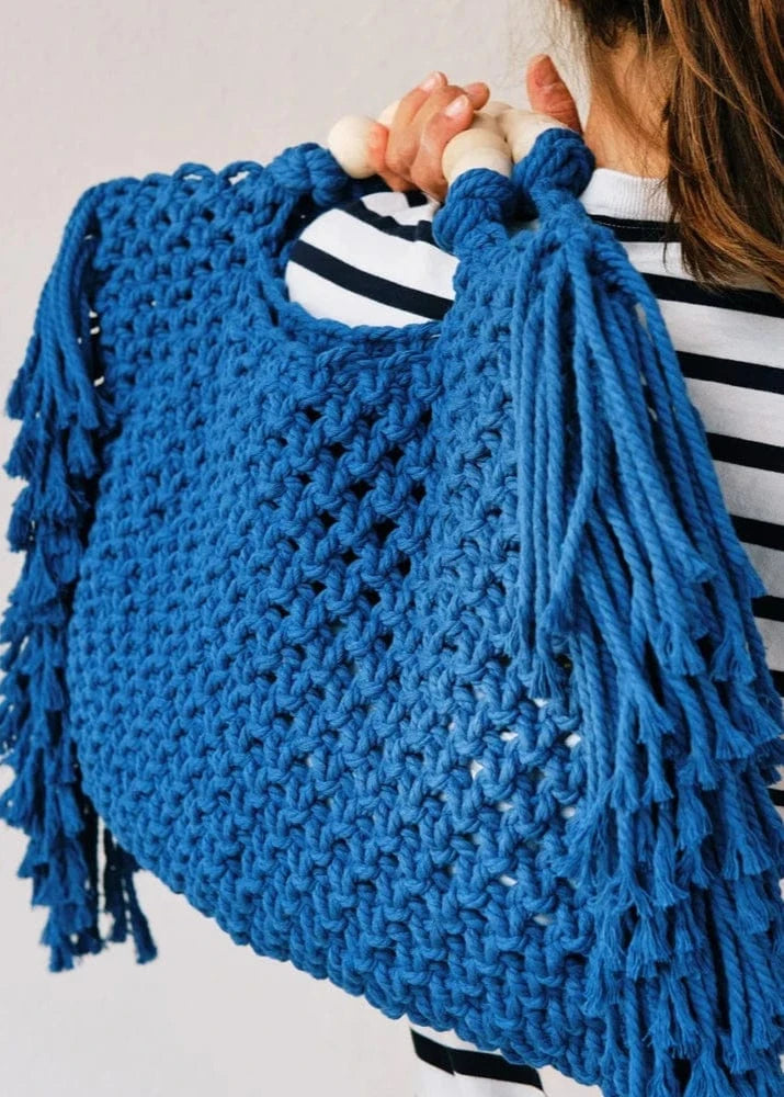 Blue knitted bag with tassels held by a person wearing a striped shirt.