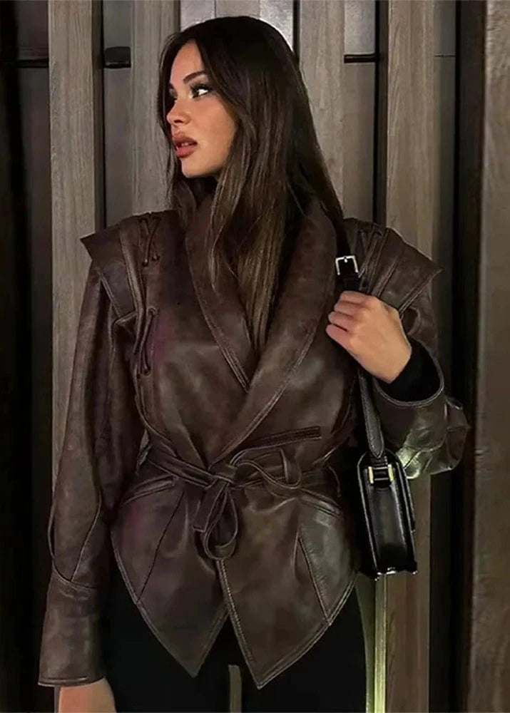 Woman wearing a brown leather jacket holding a black handbag indoors.
