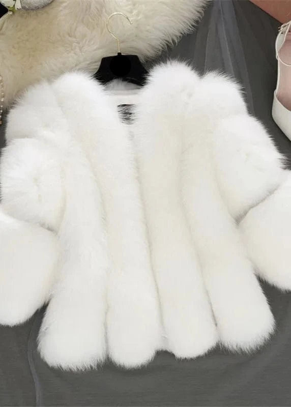 White fur coat with a black bow on a gray surface, next to white high heels and jewelry.