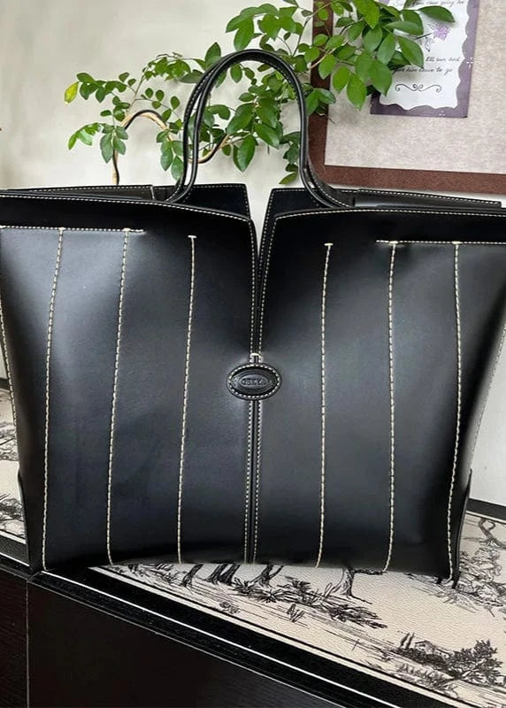 Black leather tote bag with white stitching on a decorative surface with plants in the background