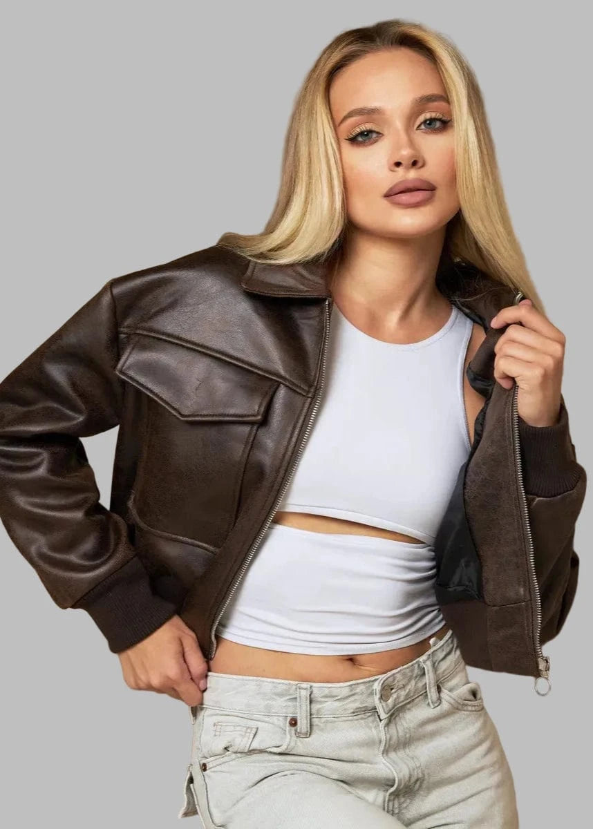 Woman wearing a brown leather jacket, white top, and light blue jeans against a beige curtain background