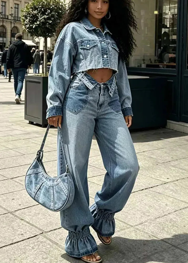 A woman walking on a city sidewalk wearing a light-wash blue denim outfit consisting of a cropped long-sleeve denim jacket and matching wide-leg ruffled jeans, carrying a light blue denim crescent shoulder bag.