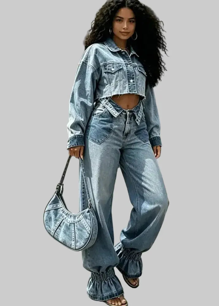 A woman stands on a city sidewalk wearing a light-wash blue denim outfit consisting of a cropped denim jacket and matching wide-leg jeans with gathered cuffs. She carries a matching blue denim crescent shoulder bag.