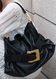 A black slouchy shoulder bag with a wide strap and a large gold buckle, worn by a person in a white knit sweater against a gray textured wall.