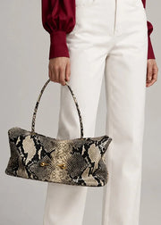 A person wearing white trousers and a burgundy blouse holds a snakeskin-patterned handbag with a top handle against a light gray studio background.
