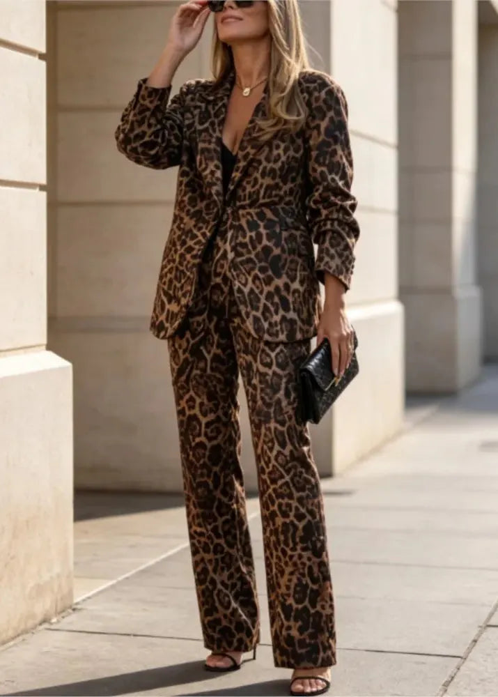 A woman wearing a leopard print blazer and matching wide-leg pants, standing outdoors on a sidewalk near beige stone columns. She is holding a small black clutch and wearing sunglasses.
