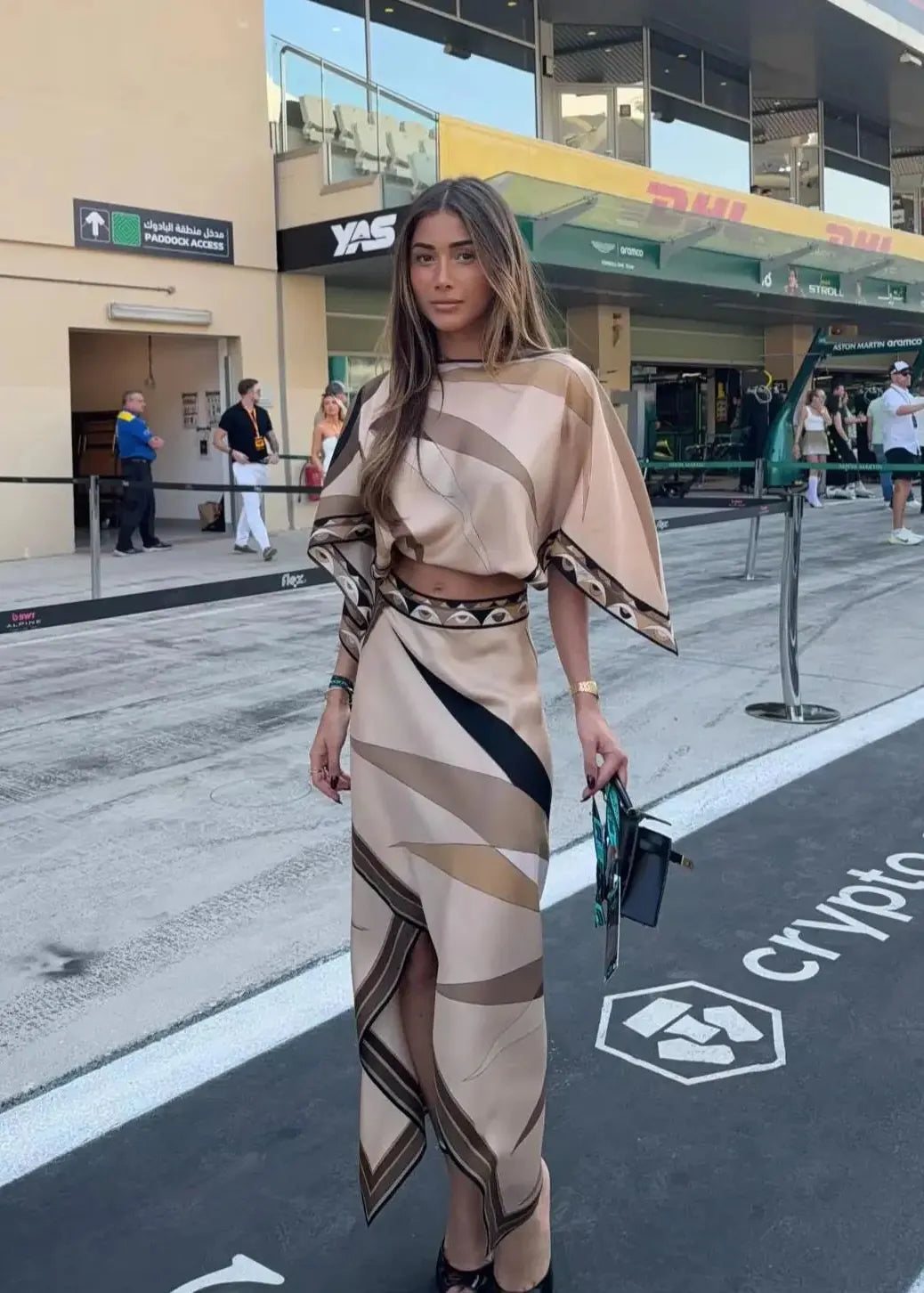 A woman stands outdoors on a street or event area wearing a beige and brown geometric print two-piece outfit with a cropped top and matching skirt. She is holding a small black handbag, with buildings, signage, and other people visible in the background.