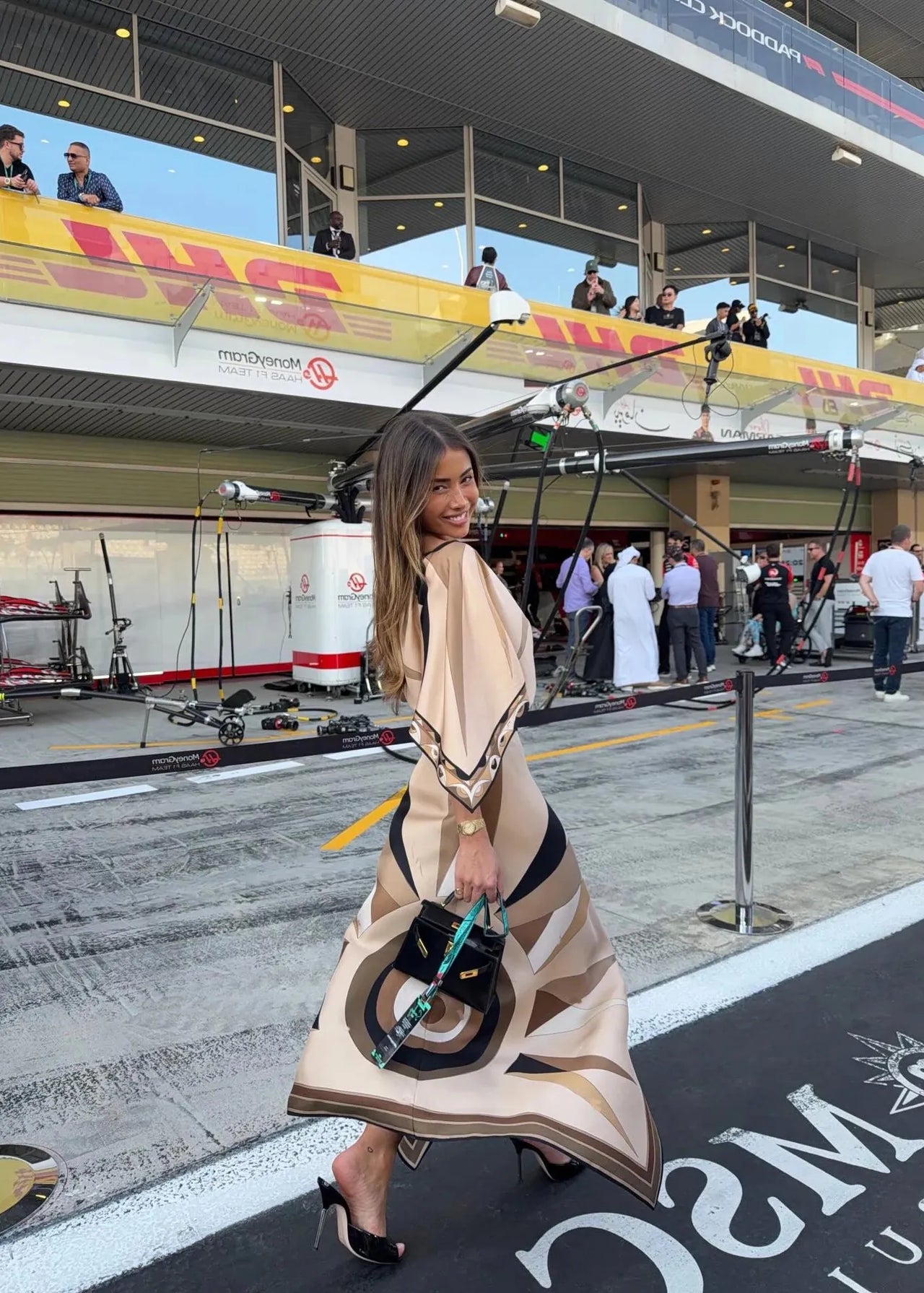 Woman in a stylish outfit with a large umbrella at a race track, with 'DHL' and 'Maserati' branding visible.