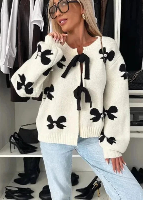 A woman wearing a black and multicolor loose-fitting sweater with a round neckline and long sleeves, paired with light blue jeans. She is also wearing glasses and has her handbag in the background.