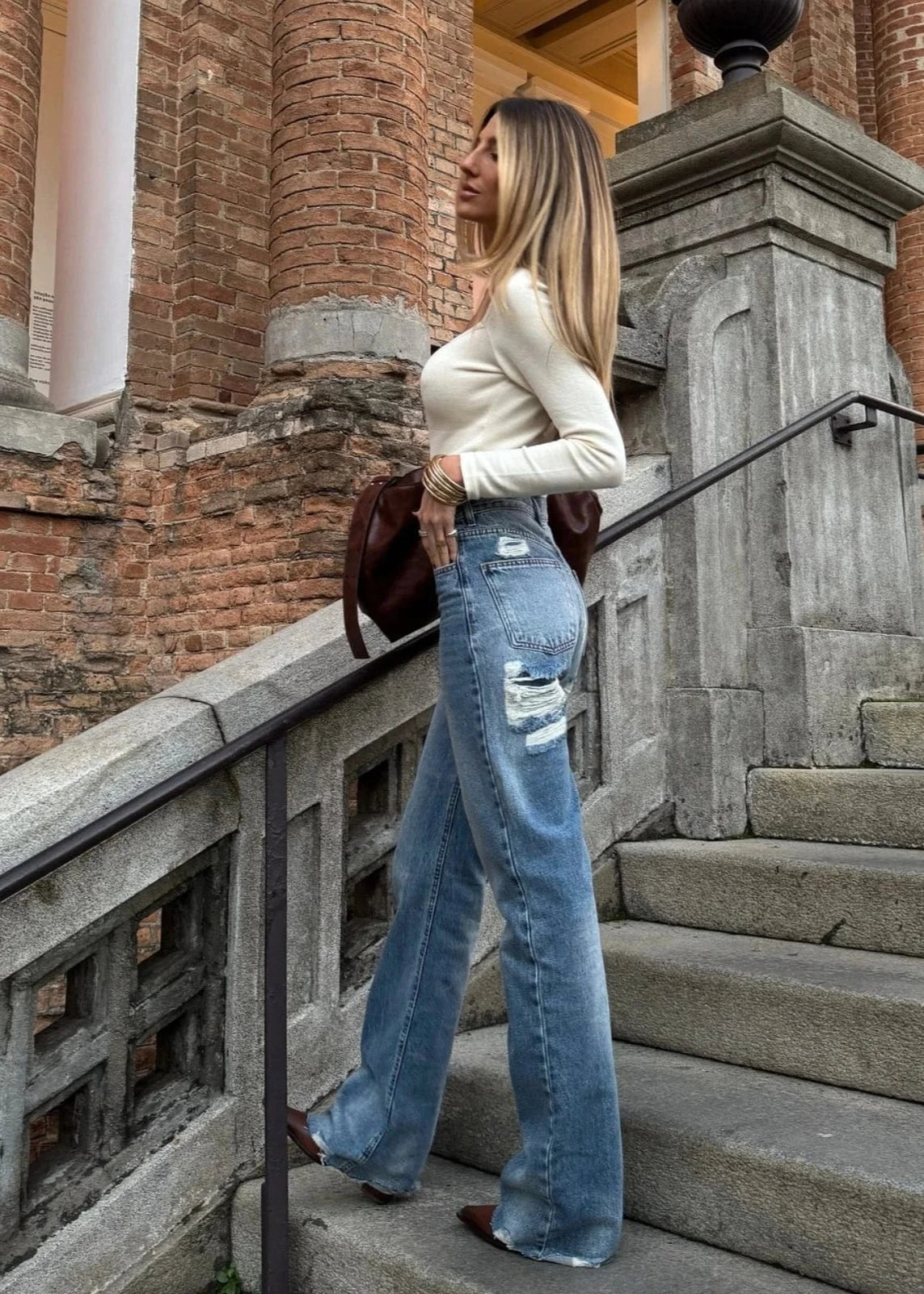 Woman in a white top and blue jeans standing on stone steps with a brick building in the background