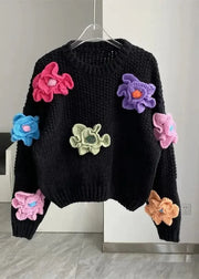 Black sweater with colorful flower patches on a hanger against a neutral wall.