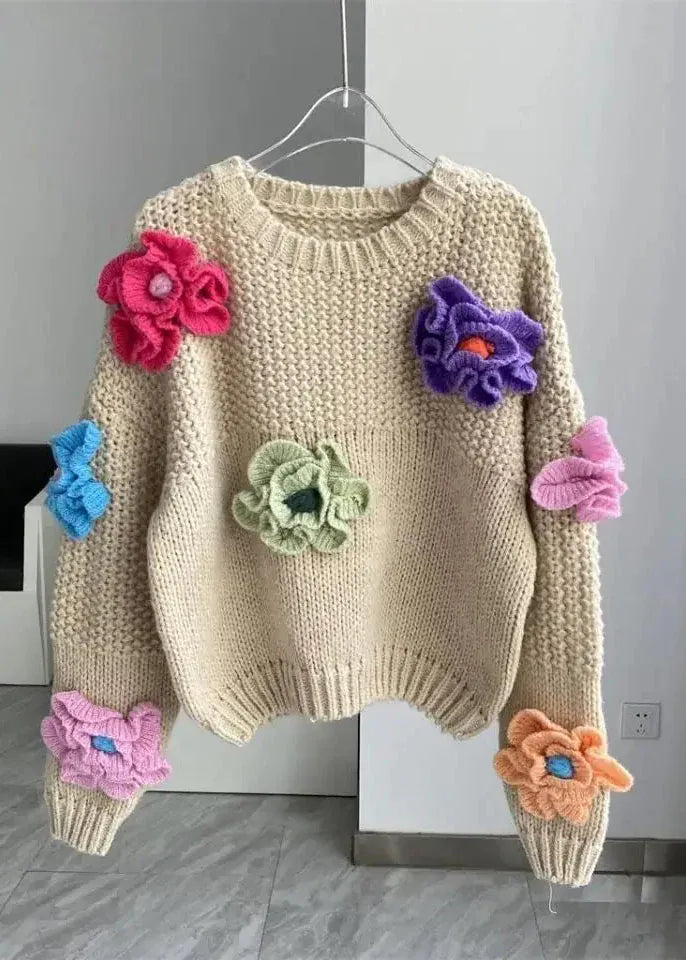 Beige sweater with colorful flower appliqués on a hanger against a gray wall.
