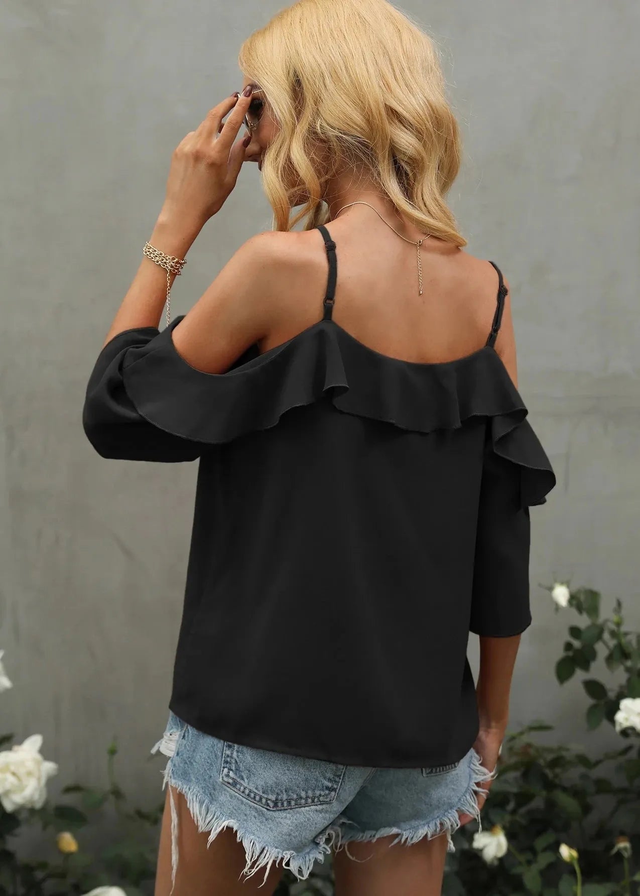Woman wearing a black ruffled top and denim shorts against a gray wall with white flowers.