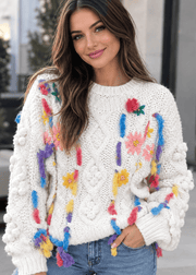 A woman wearing a cream-colored chunky knit sweater decorated with colorful yarn flowers and tassels, paired with blue jeans.