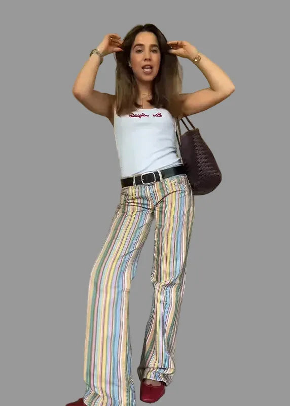 Woman wearing a white tank top and colorful striped pants, holding a purple handbag indoors.