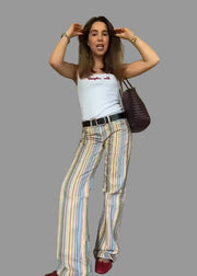 Woman wearing a white tank top and colorful striped pants, holding a purple handbag indoors.