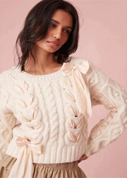 Woman wearing a cream sweater with bow details against a pink background