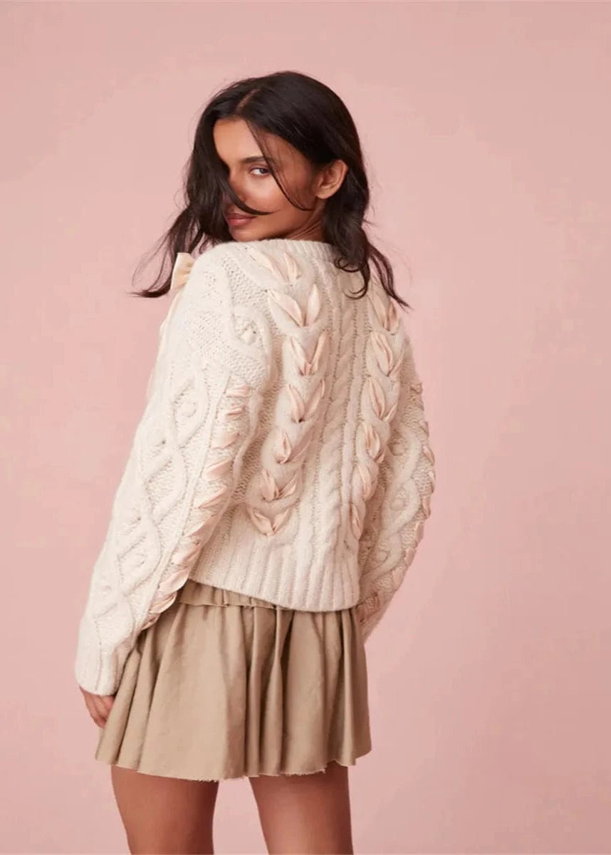 Woman wearing a cream cable knit sweater and beige skirt on a pink background