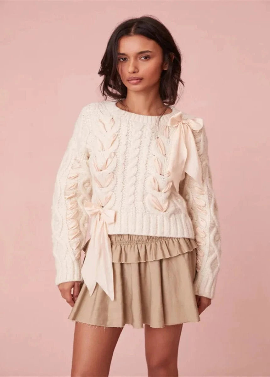 Woman wearing a cream cable knit sweater and beige skirt against a pink background