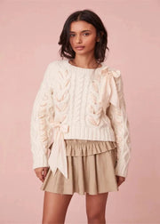 Woman wearing a cream cable knit sweater and beige skirt against a pink background