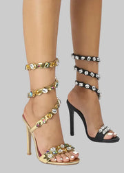 Gold and black high-heeled sandals with embellishments on a white background