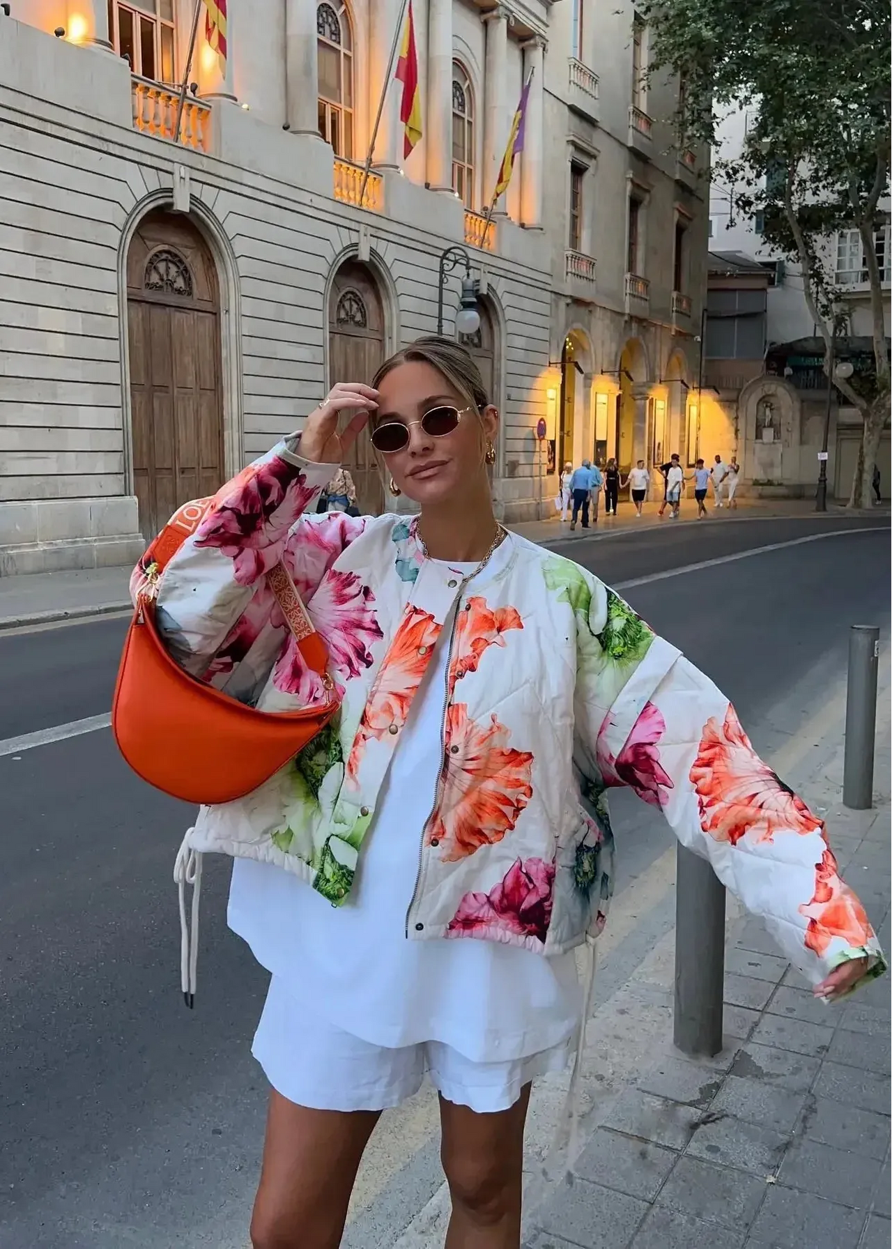 Woman in a floral jacket and white shorts holding an orange bucket hat on a city street.