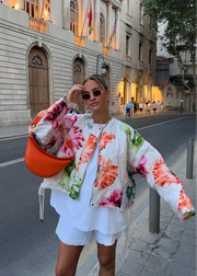 Woman in a floral jacket and white shorts holding an orange bucket hat on a city street.