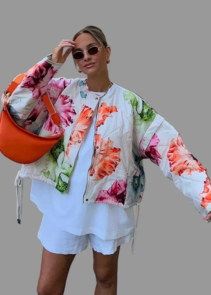 Woman in a floral jacket and white shorts holding an orange bag on a city street.