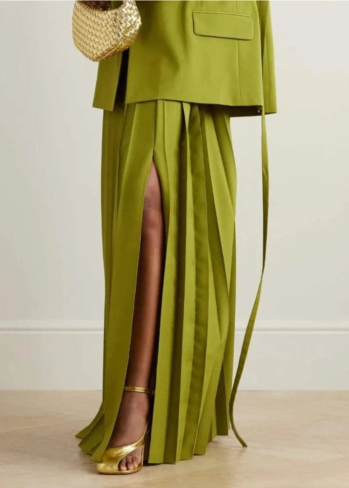 Green outfit with a skirt and jacket, gold sandals, and a woven bag on a light wooden floor.