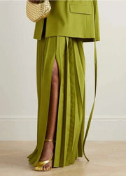 Green outfit with a skirt and jacket, gold sandals, and a woven bag on a light wooden floor.