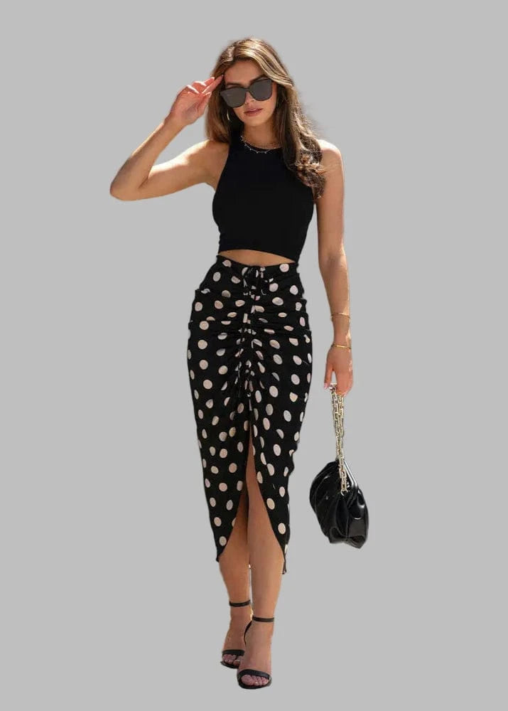 Woman wearing a black top and polka dot skirt with sunglasses and a handbag, standing against a gray wall.