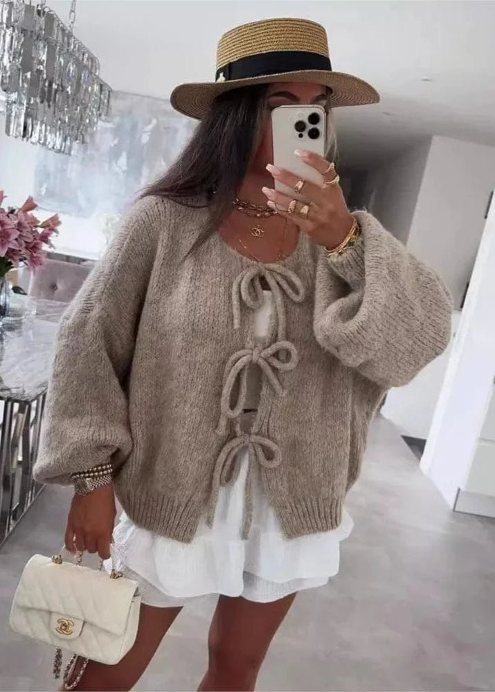 Woman wearing a beige knitted cardigan with bow details, white dress, and straw hat indoors.