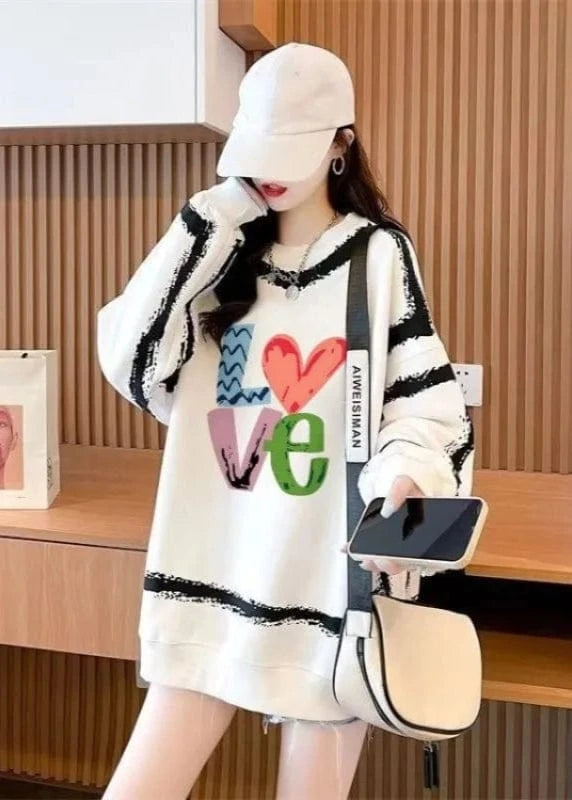 Person wearing a white sweater with colorful 'LOVE' design, holding a phone and a beige bag in an indoor setting.