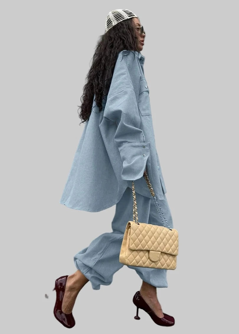 Woman in a light blue outfit holding a beige handbag against a gray wall.