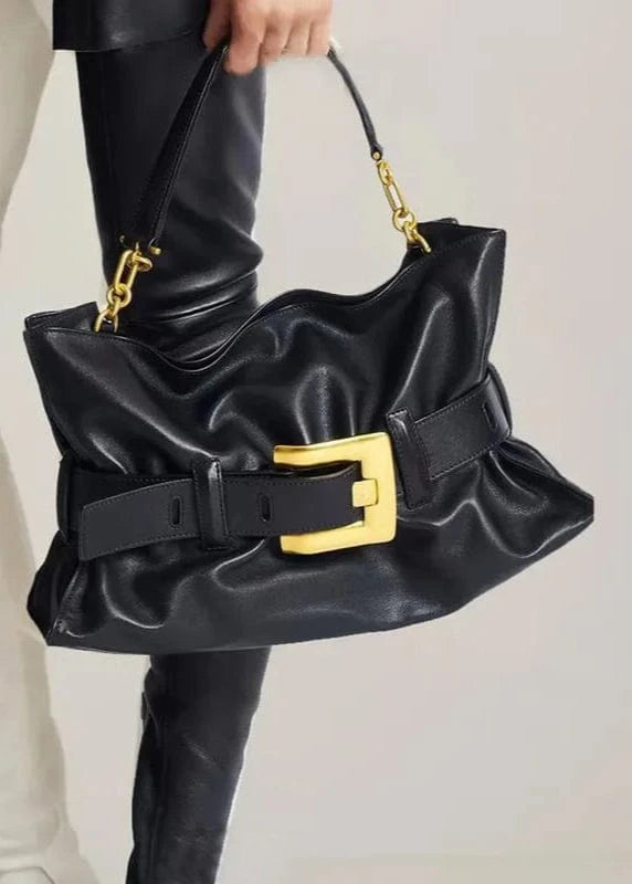 Black handbag with gold buckle held by a person wearing white pants.