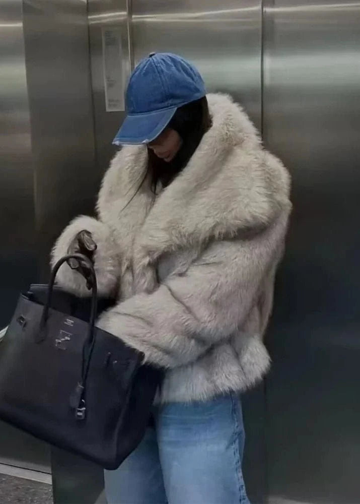 Person wearing a fluffy white coat and blue cap holding a black handbag in an elevator.