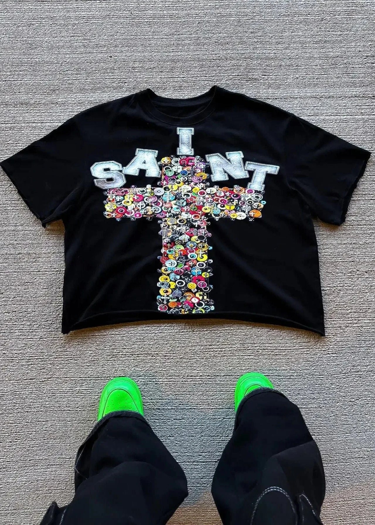 Black t-shirt with colorful cross design and 'I SAINT' text on a gray floor.