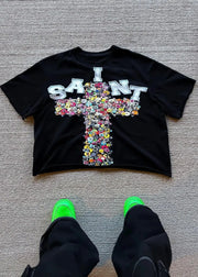 Black t-shirt with colorful cross design and 'I SAINT' text on a gray floor.