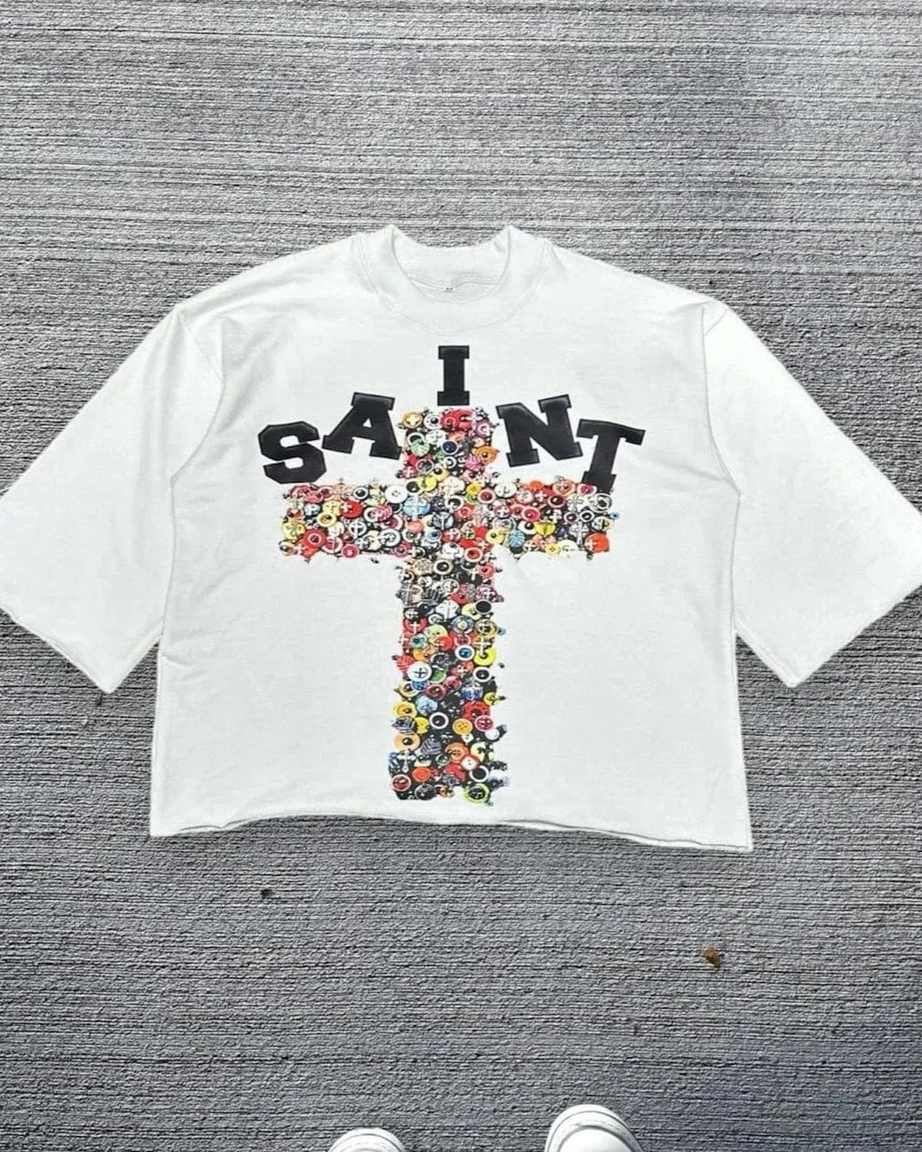 White t-shirt with floral cross design and 'I SAINT' text on a gray textured background