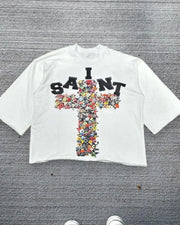 White t-shirt with floral cross design and 'I SAINT' text on a gray textured background