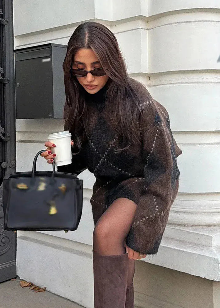 Woman holding a black handbag and a coffee cup, standing against a white wall.