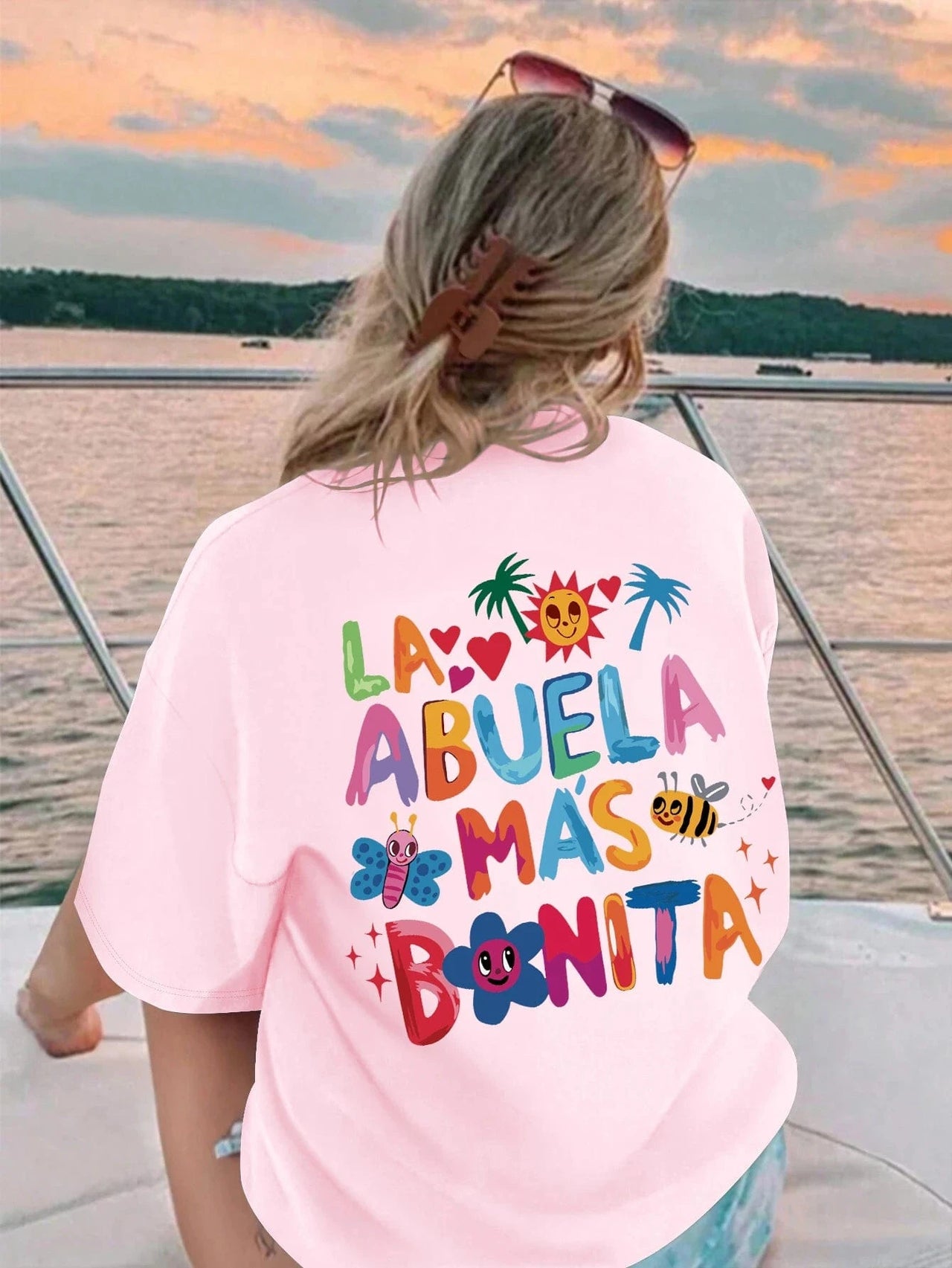 A person wearing a white crew neck t-shirt with the text 'LA ABUELA MAS BONITA' printed on the back, in a colorful font, accompanied by decorative symbols like hearts and flowers.