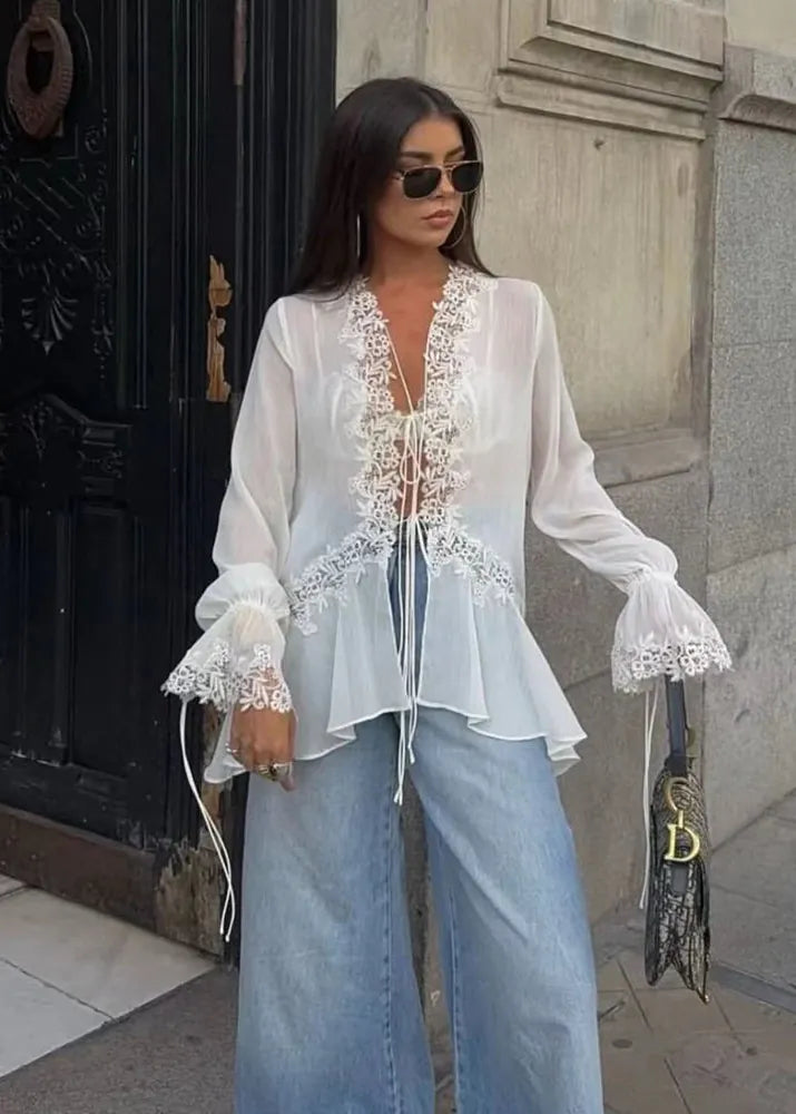 Woman wearing a white lace blouse and blue jeans standing against a building.
