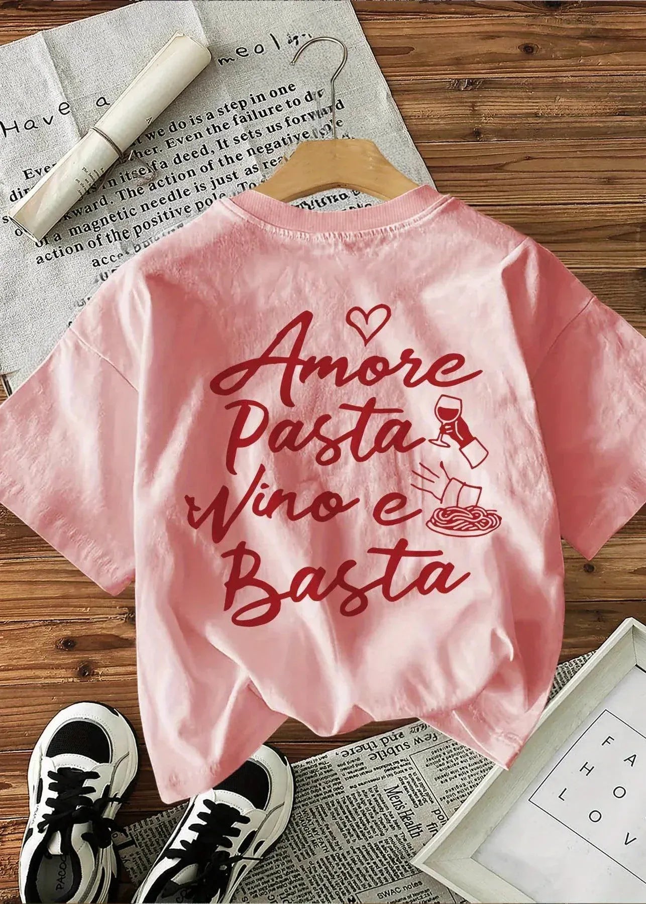 Pink t-shirt with 'Amore Pasta Wino e Basta' text on a wooden surface with newspaper and shoes.