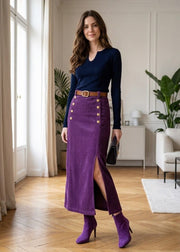 A woman models a long purple ribbed skirt with gold buttons and a front slit, paired with a navy long-sleeve top, brown belt, and purple heeled ankle boots in a bright room with wood floors.